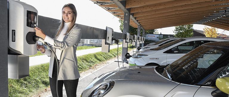 Eine Frau mit Blazer lacht in die Kamera und löst gerade einen Ladevorgang an einer Ladestation aus. Im Hintergrund ist eine Flotte von Elektrofahrzeugen zu sehen 