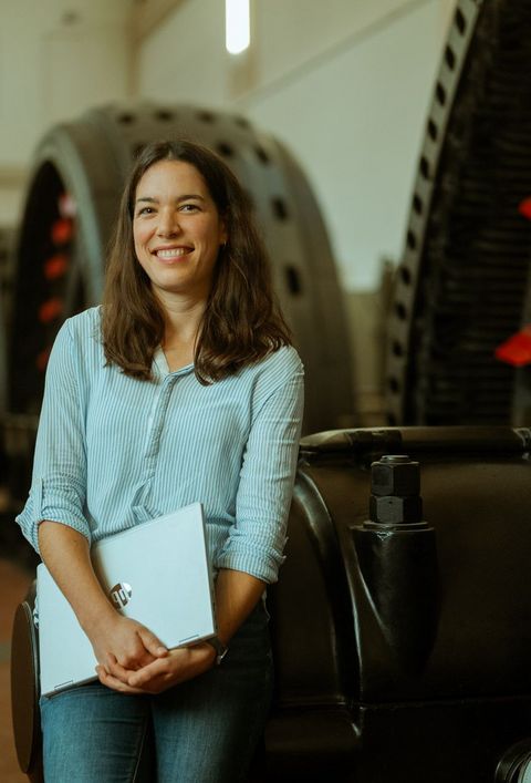 Das Bild zeigt eine Person mit Laptop in der Hand. Im Hintergrund sind große Räder zu sehen, die das Wasserkraftwerk betreiben. 