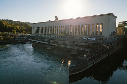 Wasserkraftwerk an einem Fluss mit Maschinenhalle und Wehranlage.