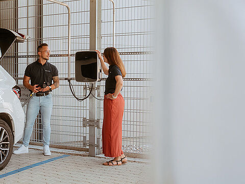Weißes Elektroauto steht an einer Wandladestation, zwei Personen daneben.
