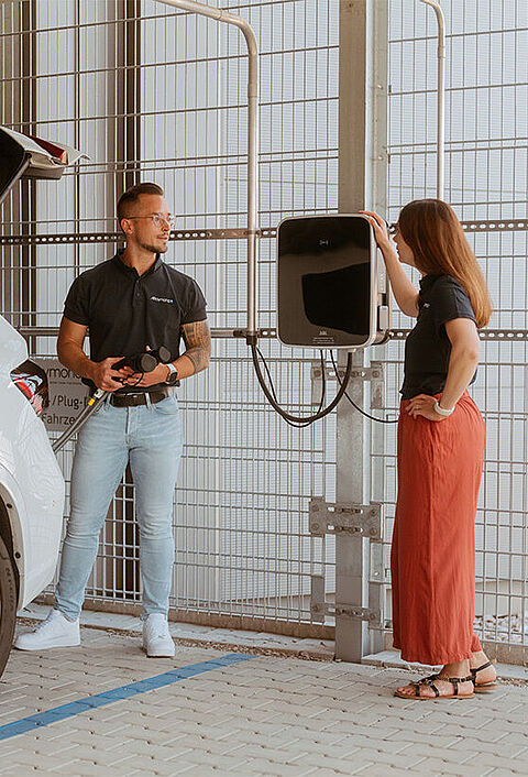 Weißes Elektroauto steht an einer Wandladestation, zwei Personen daneben.