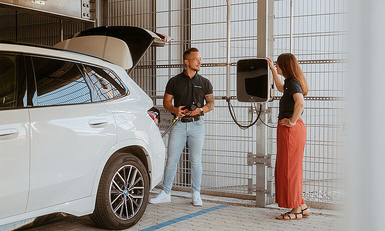 Weißes Elektroauto steht an einer Wandladestation, zwei Personen daneben.