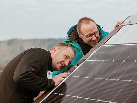 Zwei Personen arbeiten an einem geneigten Solarpanel, im Hintergrund eine hügelige Landschaft.