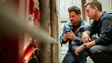 Zwei Personen inspizieren eine Maschine, eine davon dokumentiert mit dem Smartphone.