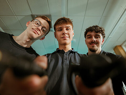 Drei Personen halten technische Geräte in der Hand in einem Innenraum in Richtung Kamera.