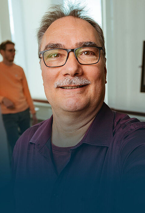 Ein älterer Mann macht ein Selfie in einem Büro. Im Hintergrund sieht man drei seiner Kollegen arbeiten.