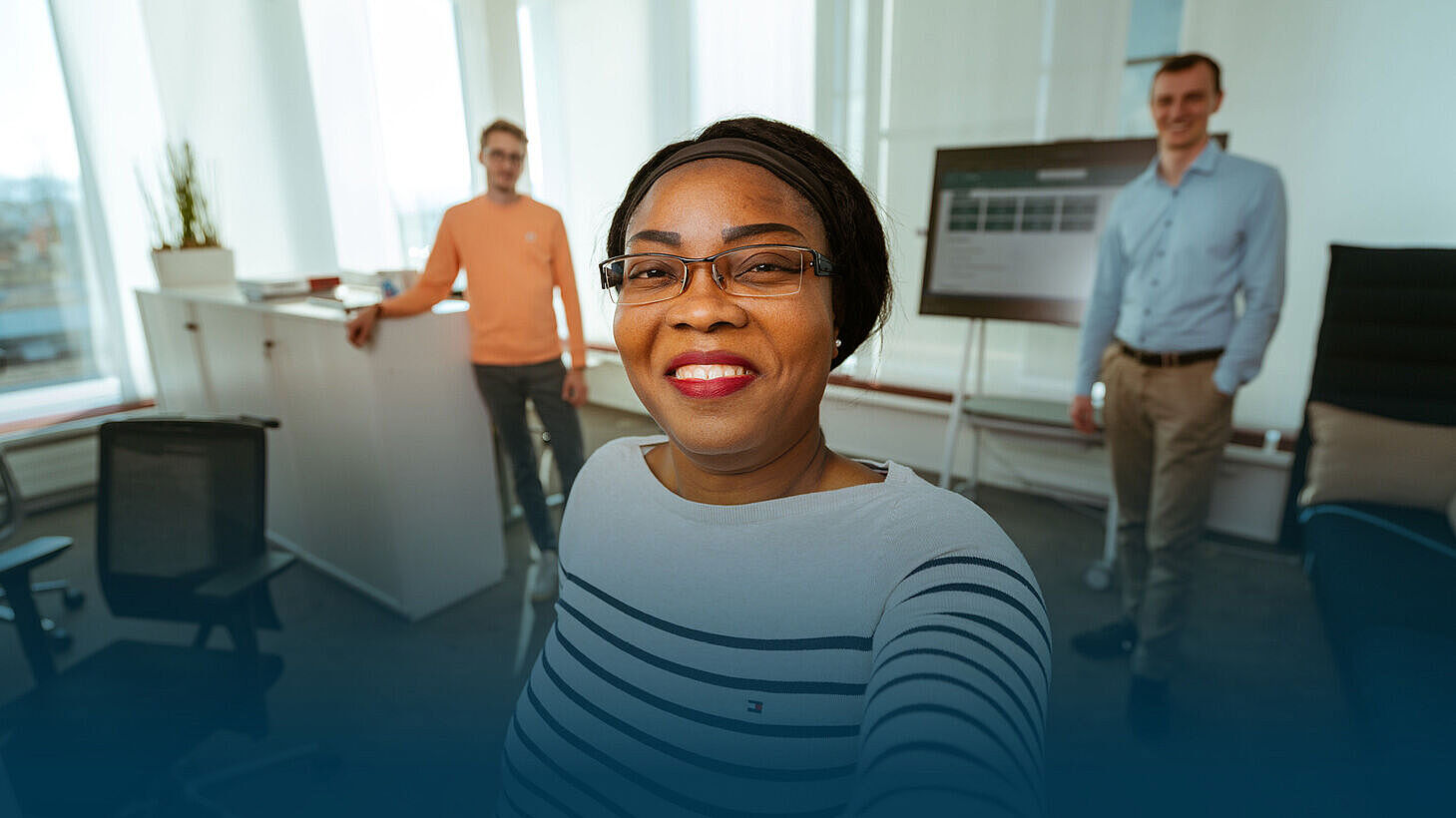 Eine Frau mit Brille macht ein Selfie in einem Büro. Im Hintergrund sieht man zwei ihrer Kollegen.
