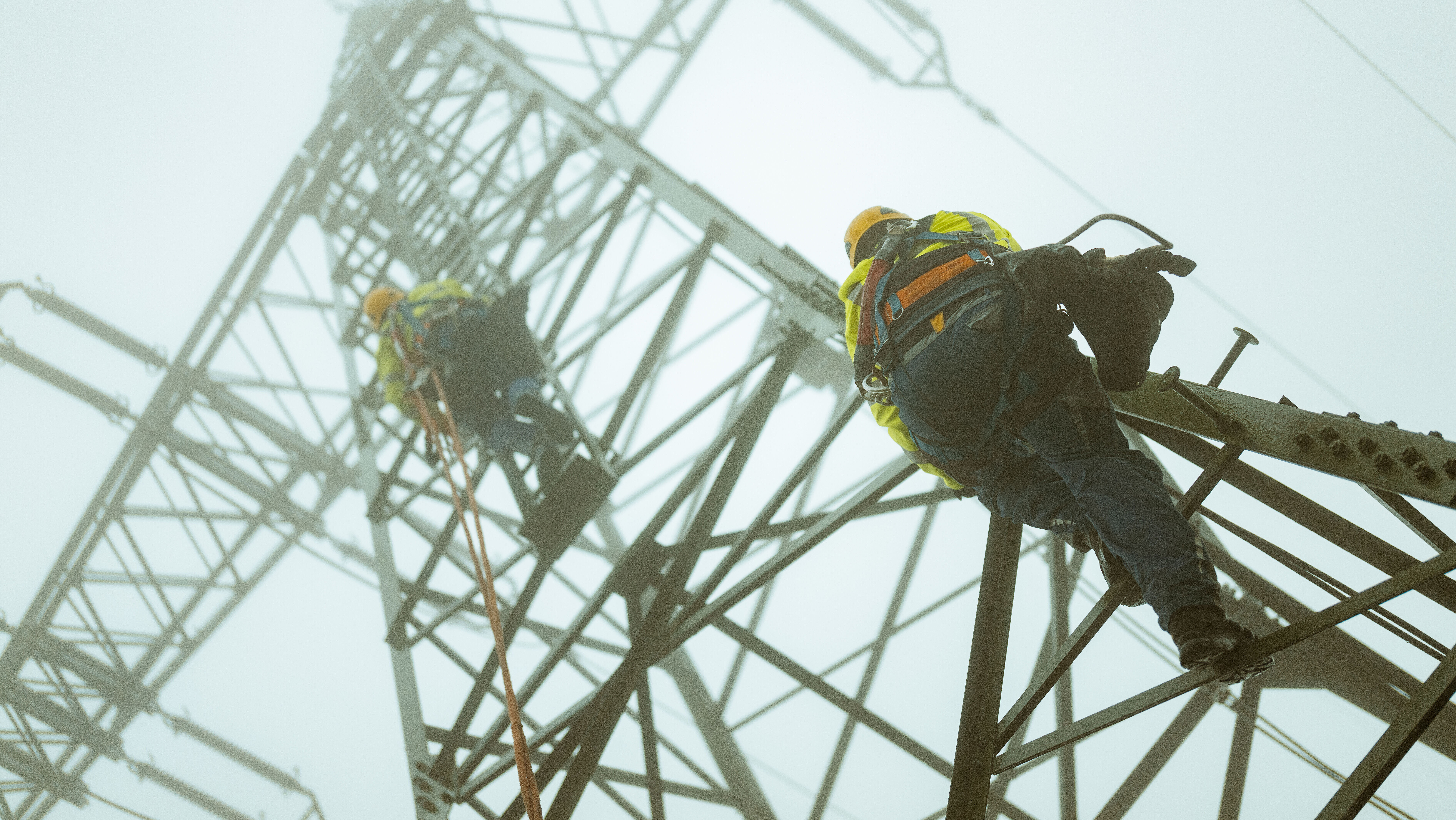 Zwei Techniker mit Sicherheitsausrüstung klettern an einem Hochspannungsmast in nebliger Umgebung.