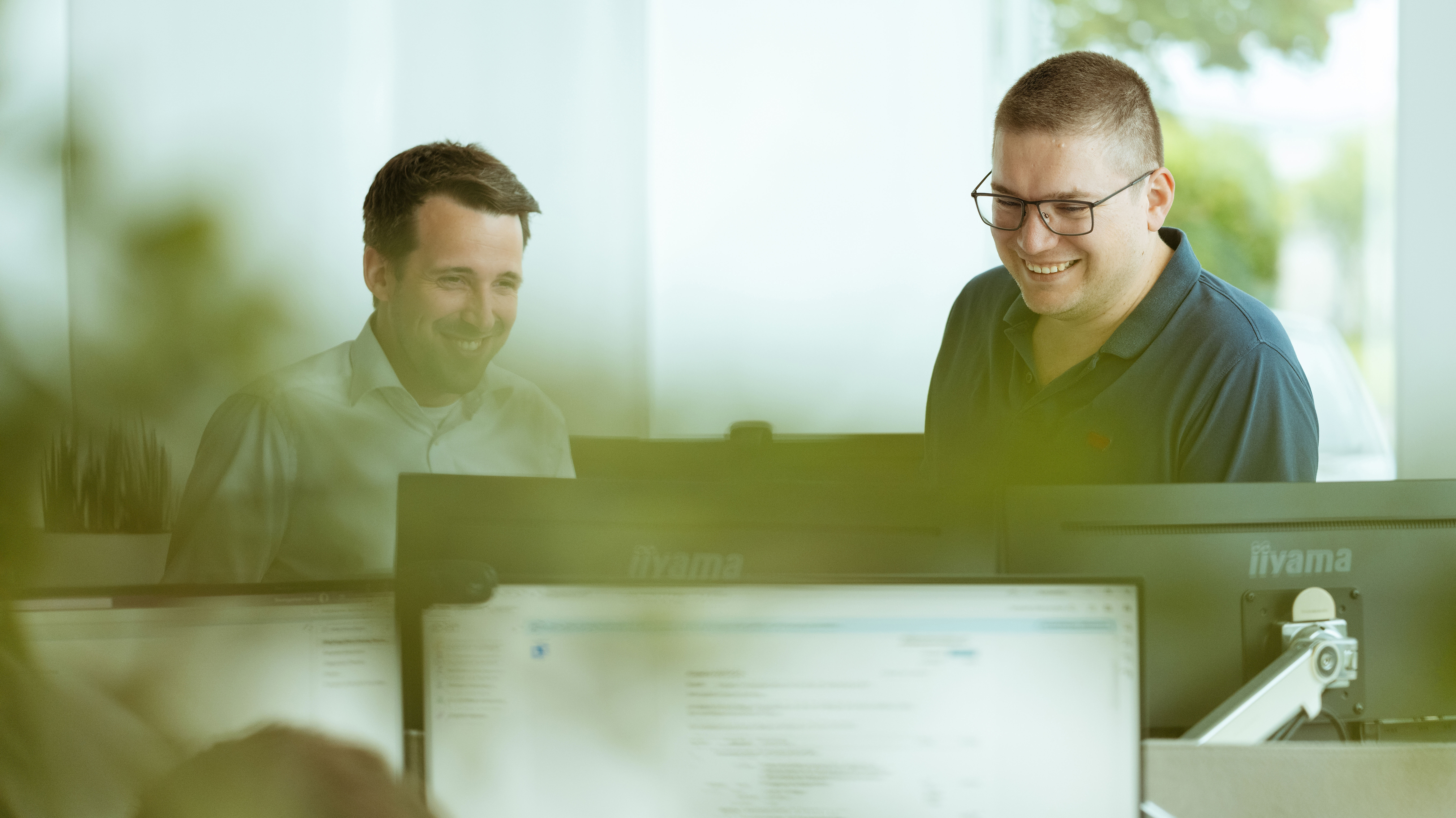 Zwei Mitarbeiter sitzen im Büro vor mehreren Monitoren und arbeiten gemeinsam an einem Projekt. Im Vordergrund ist ein weiterer Monitor und Grün zu sehen.