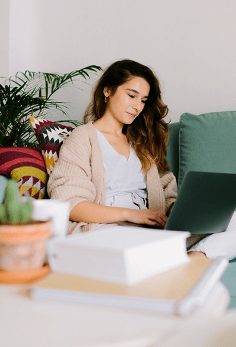 Person arbeitet mit Laptop auf Sofa, Tisch mit Büchern und Pflanze davor