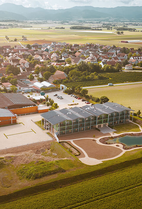 Luftaufnahme eines Gewerbegebiets mit Hallen, Parkflächen und umliegender Landschaft