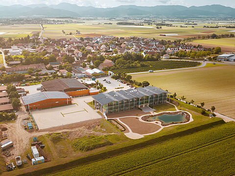 Luftaufnahme eines Gewerbegebiets mit Hallen, Parkflächen und umliegender Landschaft