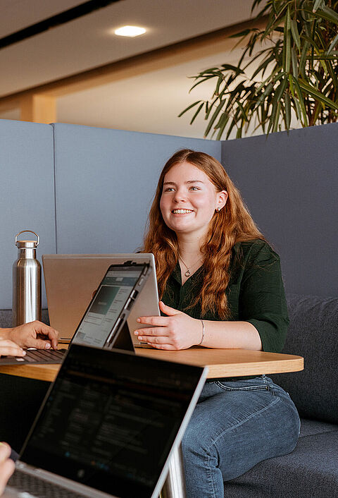 Drei Personen arbeiten gemeinsam an einem Tisch mit Laptops in einem Bürobereich.