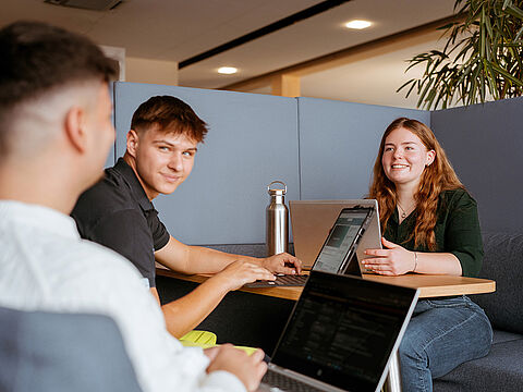Drei Personen arbeiten gemeinsam an einem Tisch mit Laptops in einem Bürobereich.