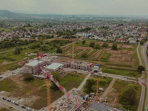 Luftaufnahme einer Baustelle mit Kränen, Gebäuden im Bau und umliegender Stadtlandschaft.