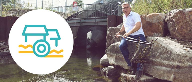 Auf dem Bild ist ein Wasserkraftwerk zu sehen, ein Mann sitzt seitlich auf einer Steinmauer und hat einen Käscher in der Hand mit welchem er Stöcke aus dem Wasser fischt. auf der linken Seite ist groß ein Icon zu sehen welches Wasserkraftwerke veranschaulichen soll. 