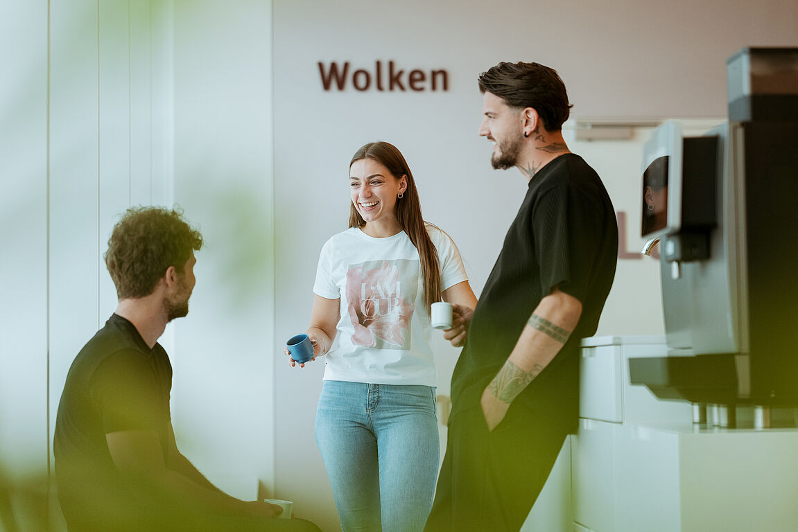 Drei Personen in einem Büro Drei Personen unterhalten sich in einem Büro neben einer Kaffeemaschine.