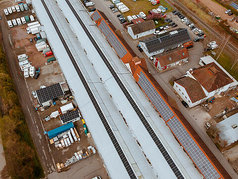 Luftaufnahme eines Industriegebiets mit langen Lagerhallen, die Solarpaneele auf den Dächern haben. Rund um die Hallen stehen viele Fahrzeuge und Container.