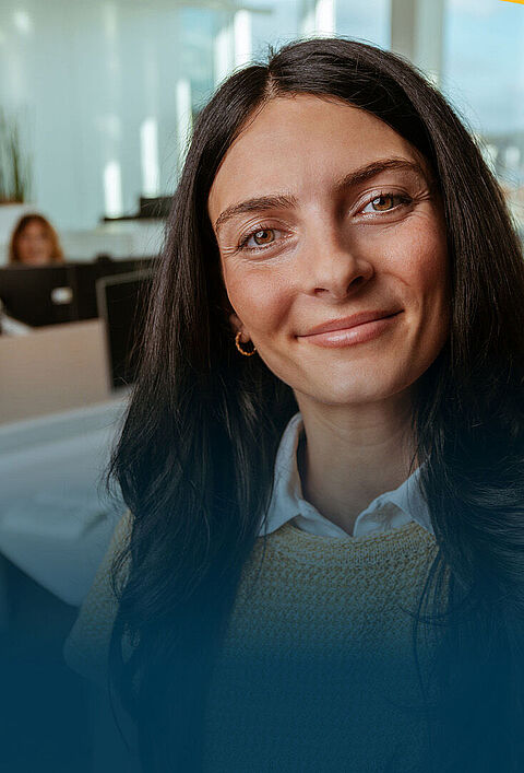 Eine Frau macht ein Selfie in einem Büro, während im Hintergrund mehrere Kolleginnen und Kollegen an ihren Arbeitsplätzen zu sehen sind.