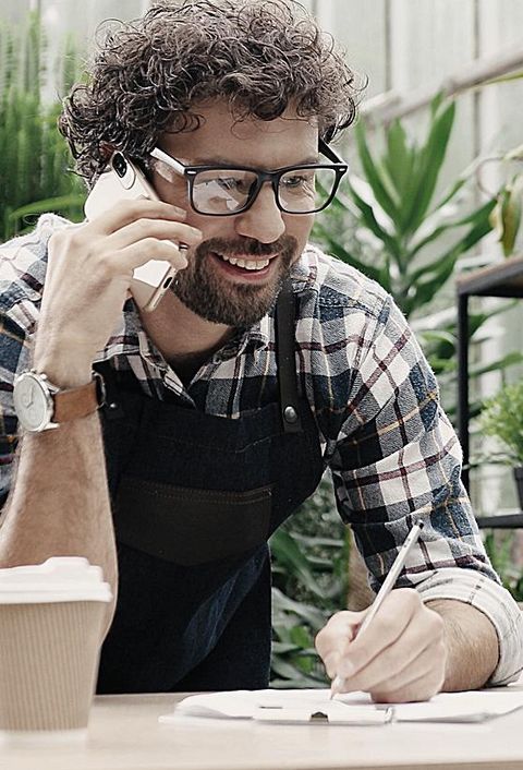 Fröhlicher männlicher Florist, der bei der Arbeit übers Smartphone telefoniert.