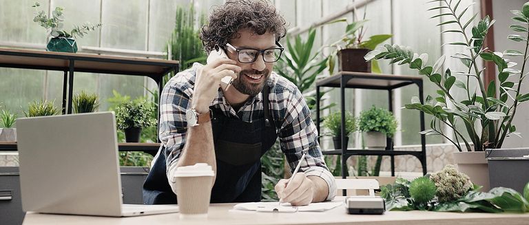 Fröhlicher männlicher Florist, der bei der Arbeit übers Smartphone telefoniert.