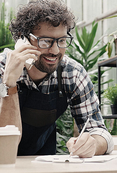 Fröhlicher männlicher Florist, der bei der Arbeit übers Smartphone telefoniert.