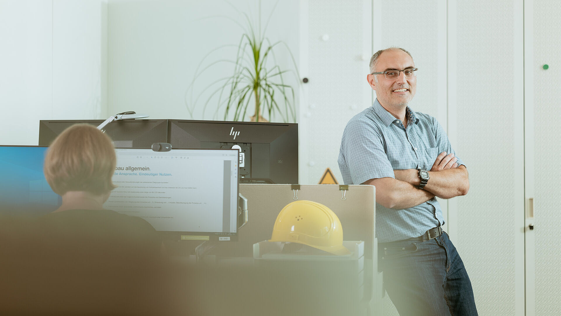 Das Bild zeigt eine Person im Büro, die sich an einem Schreibtisch anlehnt. Die Person lächelt in die Kamera . Im Vordergrund unscharf zu sehen, sieht man zwei Hinterköpfe von Personen, die gerade am Computer arbeiten. 