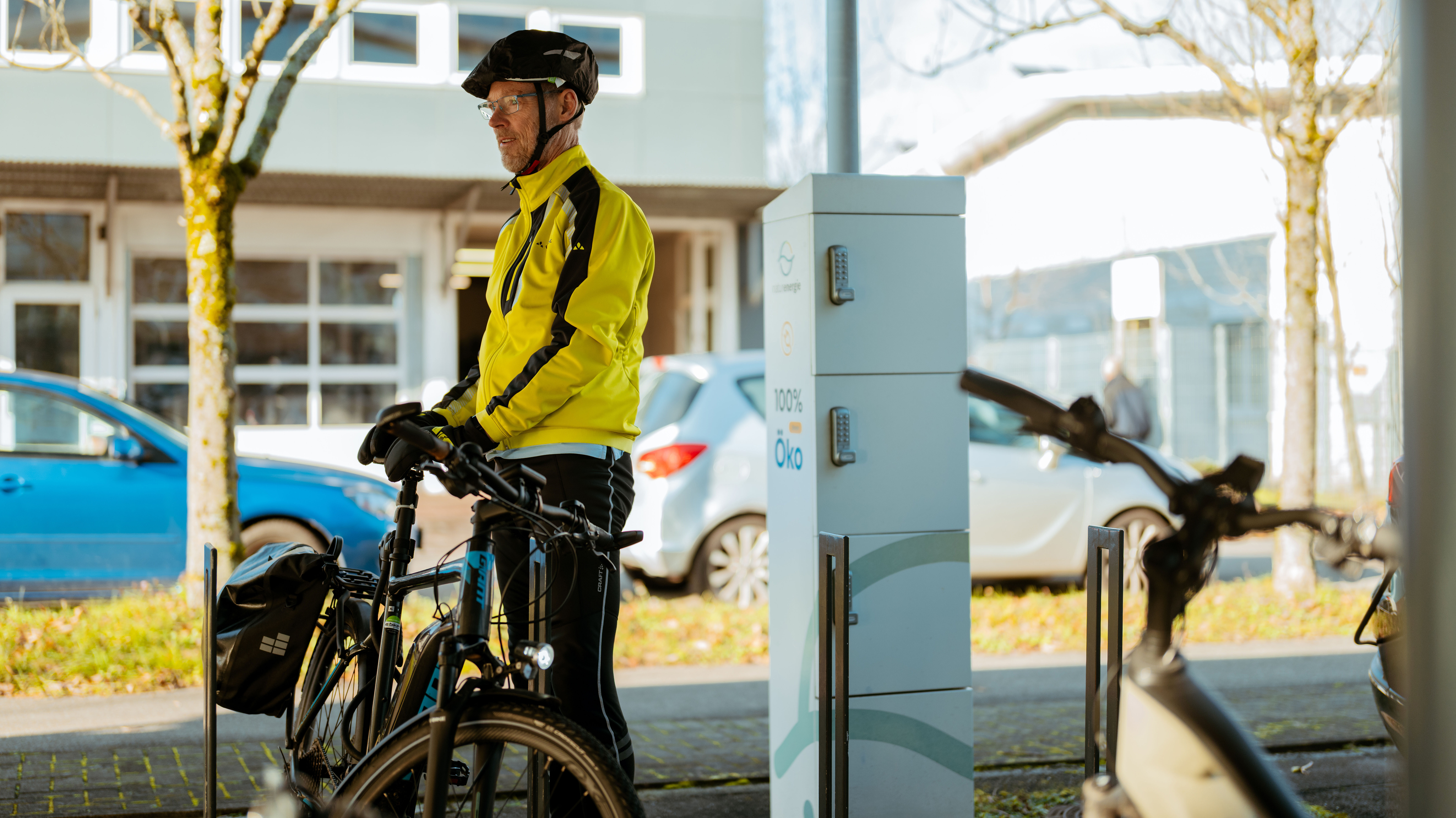Ein Mitarbeiter mit Fahrrad steht auf dem Firmenparkplatz. Neben ihm ist eine Elektroladesäule von naturenergie zu sehen.