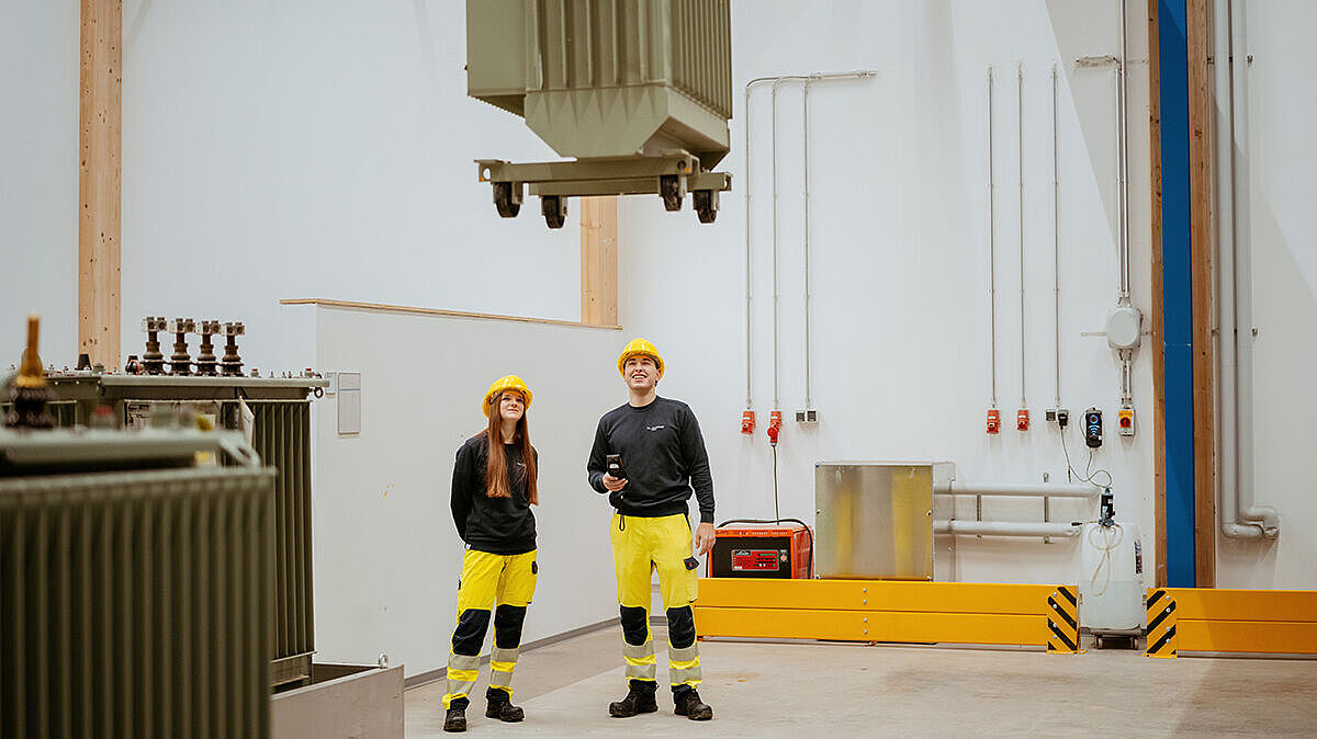 Zwei Personen stehen in einer Industriehalle unter einem angehobenen Transformator.