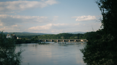 Blick über einen breiten Fluss auf ein Stauwehr, umgeben von grüner Landschaft und bewaldeten Hügeln.