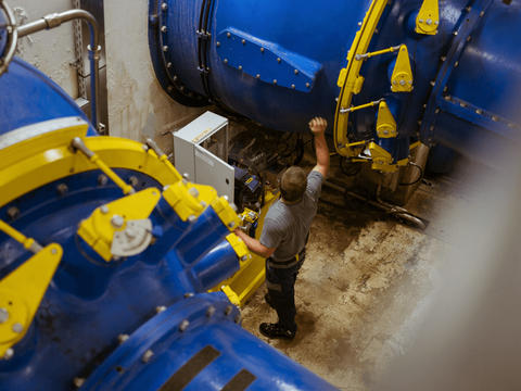 Ein Mitarbeiter steht zwischen großen blauen Industrieanlagen mit gelben Bauteilen und bedient beziehungsweise überprüft eine der Maschinen.